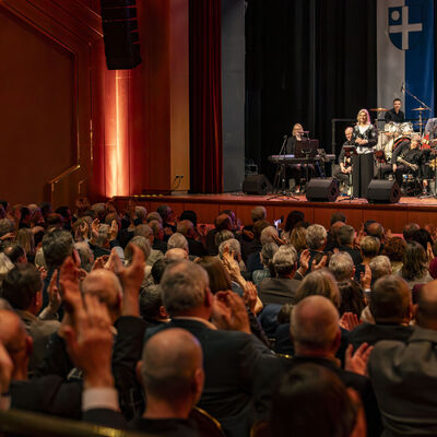 Bild vergr&ouml;&szlig;ern: Menschen sitzen im Publikum und klatschen f&uuml;r die Band auf der B&uuml;hne.
