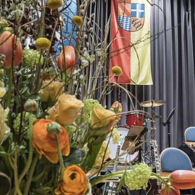 Bild vergrößern: Blumengebinde links auf der Bühne im Kurhaus Waldbronn mit Fahnen hinten des Landkreises Karlsruhe und der Gemeidne Waldbronn