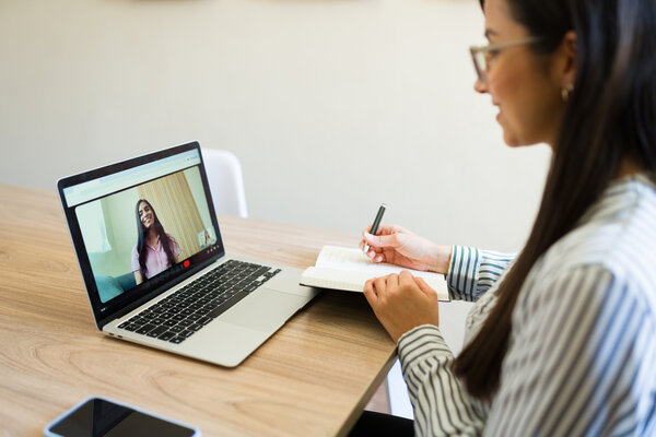 Bild vergr&ouml;&szlig;ern: Psychologin schreibt in ein Notizbuch, w�hrend sie �ber einen Laptop an einer virtuellen Therapiesitzung mit ihrer Patientin teilnimmt und ihr so ​​psychologische Fernbetreuung anbietet.